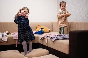 deux sœurs sont choisir vêtements de le garde-robe à Accueil sur le canapé. fille spectacles une geste avec deux des doigts. photo