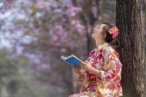 Japonais femme dans traditionnel kimono robe séance en dessous de Cerise fleur arbre tandis que en train de lire une livre pendant printemps Sakura Festival concept photo