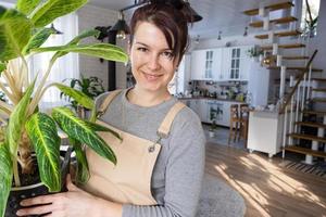 une content femme dans une vert maison avec une mis en pot plante dans sa mains sourires, prend se soucier de une fleur. le intérieur de une confortable respectueux de la nature loger, une cheminée poêle, une loisir pour croissance et reproduction plante d'intérieur photo