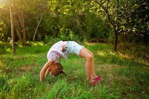 une peu fille fait du une pont, pliant sa retour dans la nature. une souple enfant, Faire gymnastique des exercices. photo