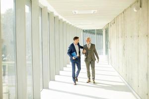 Jeune et une Sénior homme d'affaire marcher vers le bas un Bureau couloir, Profond dans conversation photo