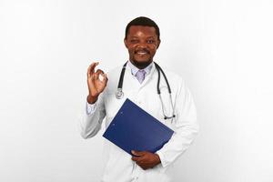 souriant noir barbu médecin homme dans blanc peignoir détient médical presse-papiers et spectacles D'accord geste photo