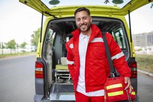 Jeune homme , une paramédical, permanent à le arrière de un ambulance, par le ouvert des portes. il est à la recherche à le caméra avec une sur de soi expression, souriant, porter une médical traumatisme sac sur le sien épaule. photo