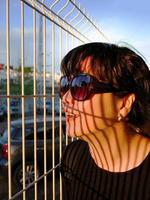 jouer de soir lumières ombre sur le affronter, portrait de une femelle asiatique dans noir robe portant des lunettes de soleil, verticale blanc métal clôture ombre moulage sur sa affronter, le coucher du soleil photo