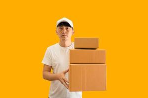 Jeune asiatique livraison homme portant casquette et blanc Vide T-shirt en portant parcelle Publier boîte isolé sur Jaune Contexte. Express livraison un service concept. photo