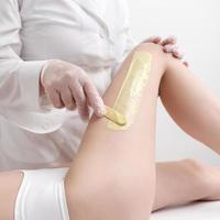 fartage procédure - appliquant vert chaud la cire sur peau jambe en utilisant spatule tandis que femme mensonge vers le bas sur canapé photo