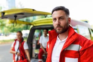 Jeune homme, paramédical permanent arrière de le ambulance. ambulanciers par le ambulance. deux ambulanciers à venir en dehors de ambulance photo