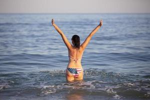 une âge moyen femme des stands dans mer l'eau avec sa bras élevé dans une maillot de bain avec un américain drapeau imprimer. photo