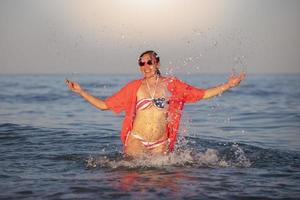 content âge moyen femme des stands dans mer l'eau avec élevé bras dans une maillot de bain avec un américain drapeau imprimer. photo