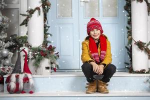 une Beau peu garçon dans brillant vêtements est assis sur le porche de une maison décoré avec Noël décorations. photo