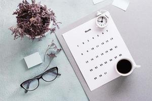 mensuel calendrier feuille, café tasse, alarme horloge, des lunettes et une bouquet de fleurs sur le tableau. temps Planification et organisation photo