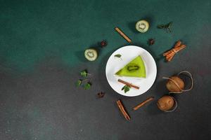 fait maison cheesecake avec kiwi dans une blanc assiette sur une vert Contexte. décortiqué avec tranche de kiwi, cannelle bâton et feuilles menthe. Haut vue photo