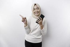femme asiatique excitée portant le hijab pointant vers l'espace de copie à côté d'elle tout en tenant son téléphone, isolée sur fond blanc photo