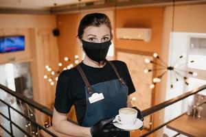 une serveuse porte un masque noir et des gants jetables sert une tasse de café dans un restaurant photo