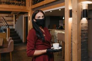 Une femme gérante de restaurant porte un masque noir et des gants jetables tenant une tasse de café dans un restaurant photo