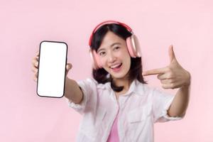 content de bonne humeur souriant asiatique femme avec sans fil écouteurs montrant Vide écran mobile téléphone ou Nouveau téléphone intelligent la musique application publicité maquette isolé sur rose studio Contexte. photo