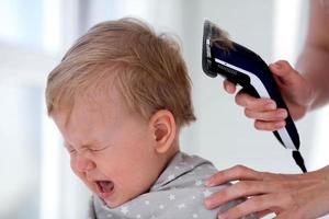 femelle mains réduire une pleurs bébé avec un électrique cheveux tondeuse dans une coiffeur. premier la Coupe de cheveux. photo