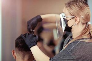 coiffeur avec Sécurité les mesures pour COVID-19 [feminine, coupes une homme social distance, Coupe cheveux avec une médical masque, visage bouclier et caoutchouc gants. photo