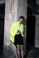 un asiatique homme dans une citron vert coloré veste penché contre une abandonné bâtiment pilier à la recherche très Beau photo