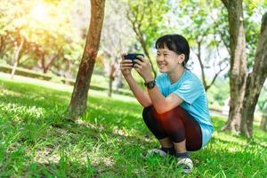 portrait de Jeune asiatique enfant fille en utilisant mobile téléphone intelligent tandis que dans le parc dans chaud printemps journée. photo