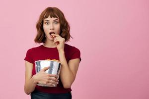 excité sous le choc mignonne roux Dame dans rouge T-shirt avec pop corn prêt pour film soir posant isolé sur plus de rose studio Contexte. copie espace bannière. mode cinéma concept. divertissement offre photo