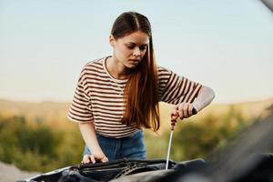 une femme voiture chauffeur ouvert le capuche cassé vers le bas le voiture tire en dehors le jauge et chèques le moteur pétrole sur le route seule. voiture réparation photo