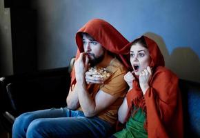 une homme et une femme avec une rouge plaid sur leur têtes sont séance sur le canapé dans de face de le la télé à l'intérieur photo