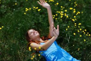 femme sourire mensonges sur le vert herbe dans une bleu été robe en dessous de le Soleil avec Jaune fleurs, bonheur, rouge longue cheveux photo