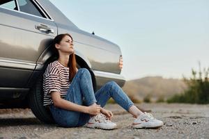 une Jeune femme est assis sur le sol près sa voiture sur le côté de le route et regards à le le coucher du soleil. camping après une difficile route voyage photo