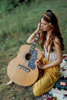 hippie femme éco amical en jouant guitare sourit et en chantant Chansons dans la nature séance sur une plaid par le Lac dans le soir dans le le coucher du soleil lumière du soleil. une mode de vie dans harmonie avec le corps et la nature photo