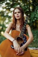 une Jeune hippie femme avec une guitare dans sa mains sourit gentiment dans le caméra sur une voyage à la nature mode de vie dans harmonie photo