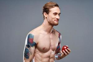 sexy athlète avec pompé en haut muscles mange un Pomme sur une gris Contexte et une tatouage sur le sien bras photo