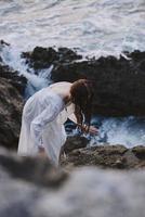 jolie femme dans blanc robe des stands sur le rochers paysage océan inchangé photo