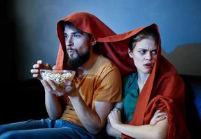 effrayé femme avec une rouge plaid sur sa tête et une homme avec une assiette de pop corn dans une foncé pièce photo