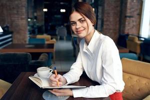 une femme dans une chemise avec une carnet et une stylo dans sa main est assis à une table dans une restaurant sur un tapissé chaise photo