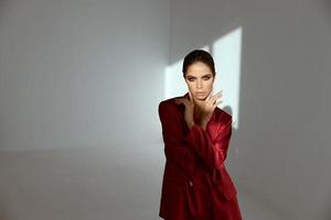 une femme avec brillant maquillage et une rouge veste dans une foncé pièce et une brillant lumière de le fenêtre photo