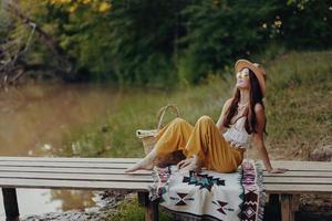 femme dans hippie éco robe est assis dans la nature par le Lac sur une pont relaxant avec une panier de nourriture et admiratif le paysage en train de regarder le le coucher du soleil photo