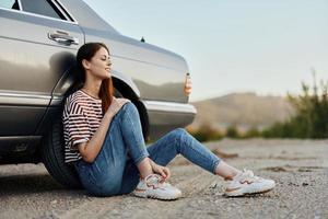 une Jeune femme est assis sur le sol près sa voiture sur le côté de le route et regards à le le coucher du soleil photo