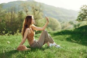 femme blogueur est assis sur le vert herbe dans une parc et prend des photos de se sur sa téléphone contre le toile de fond de une été paysage. Jeune les gens mode de vie et préoccupation pour le environnement