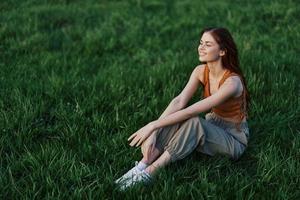 une magnifique fille relaxant dans la nature séance dans un pantalon et Haut sur le vert herbe dans le des rayons de le réglage été Soleil. le concept de santé et se soucier pour le corps et le environnement photo