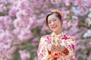 Japonais femme dans traditionnel kimono robe en portant sucré hanami dango dessert tandis que en marchant dans le parc à Cerise fleur arbre pendant printemps Sakura Festival avec copie espace photo