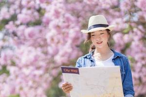 asiatique femme touristique en portant ville carte tandis que en marchant dans le parc à Cerise fleur arbre pendant printemps Sakura fleur Festival concept photo