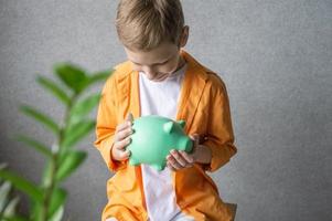 une mignonne garçon dans une chemise détient une porcin banque dans le sien mains et regards dans le porcin banque photo