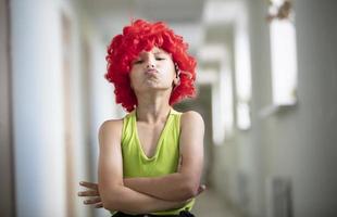 une enfant dans une brillant perruque. marrant garçon avec rouge artificiel cheveux. artiste écolier grimaces. photo