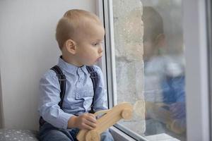 une petit enfant regards en dehors le la fenêtre. le enfant avec une jouet est assis sur une fenêtre seuil. photo