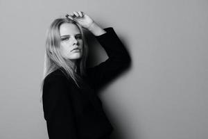 sérieux Jeune blond femme penche sur le studio mur en portant main aérien regards à caméra posant isolé dans noir veste photo