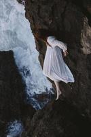 femme dans longue blanc robe humide cheveux mensonge sur une rocheux falaise été vacances concept photo