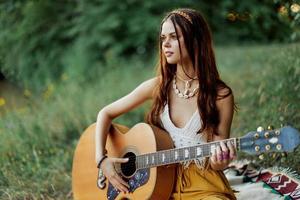 hippie femme souriant et étreindre sa guitare dans la nature dans le parc dans le le coucher du soleil lumière dans éco-vêtements photo