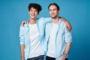 gars avec frisé cheveux dans une chemise et une T-shirt sur une bleu Contexte et une Jeune homme copains amusement photo