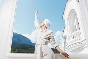 femme avec une serviette sur mon tête dans une blanc peignoir de bain rester sur le balcon dans une Hôtel parfait ensoleillé Matin photo